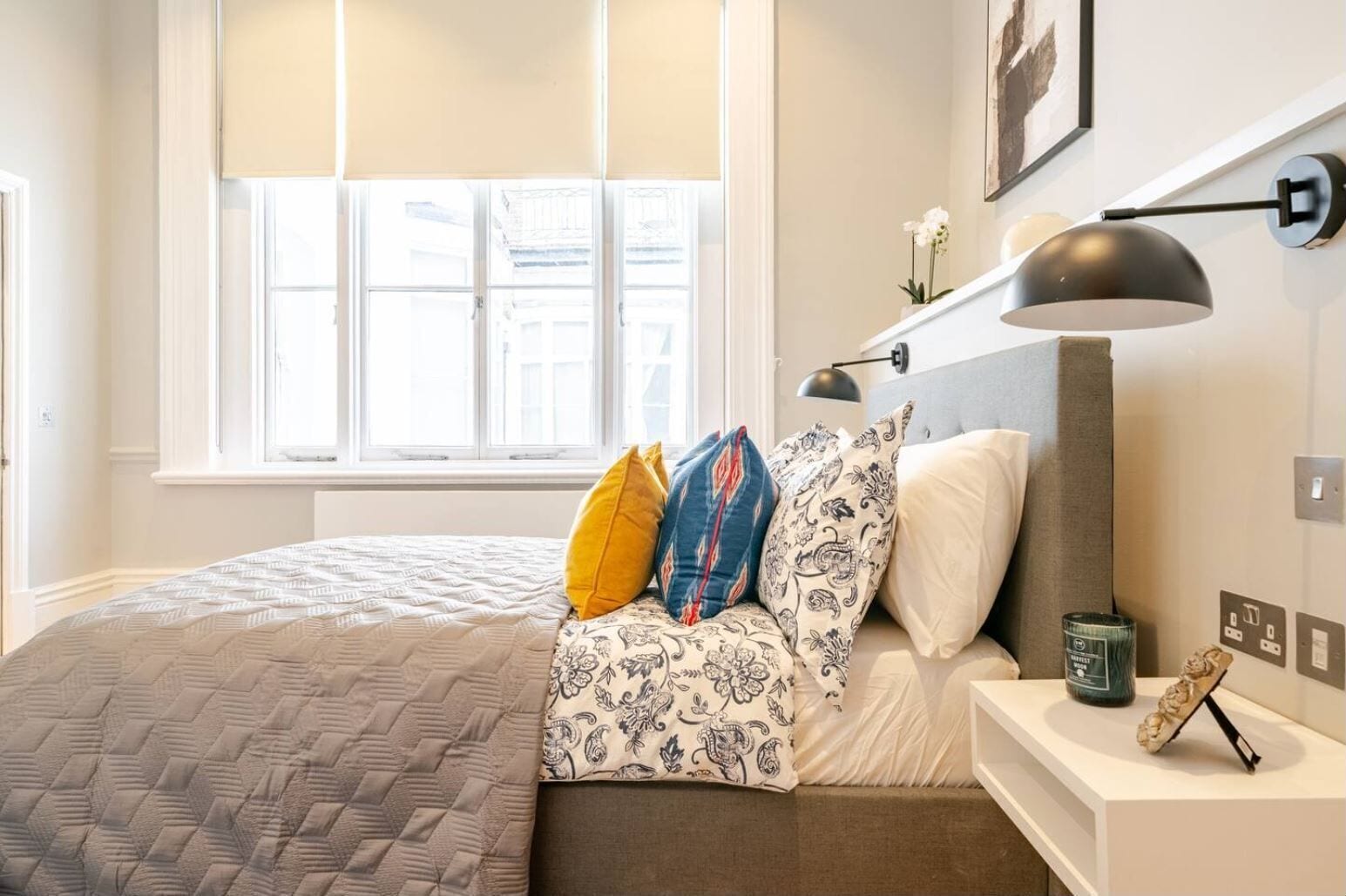 A neatly arranged bedroom with a bed, pillows, a bedside table with a lamp, and a window with blinds.