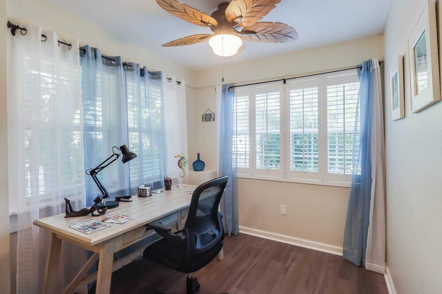 Une pièce comprenant un bureau, une chaise, un ventilateur de plafond et des fenêtres avec des rideaux.