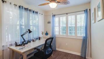 Une pièce comprenant un bureau, une chaise, un ventilateur de plafond et des fenêtres avec des rideaux.