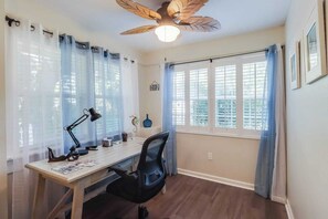 A room with a desk, chair, ceiling fan, and windows with curtains.