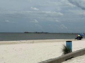 A beach with a pier extending into the water, a trash can, and a beach umbrella.