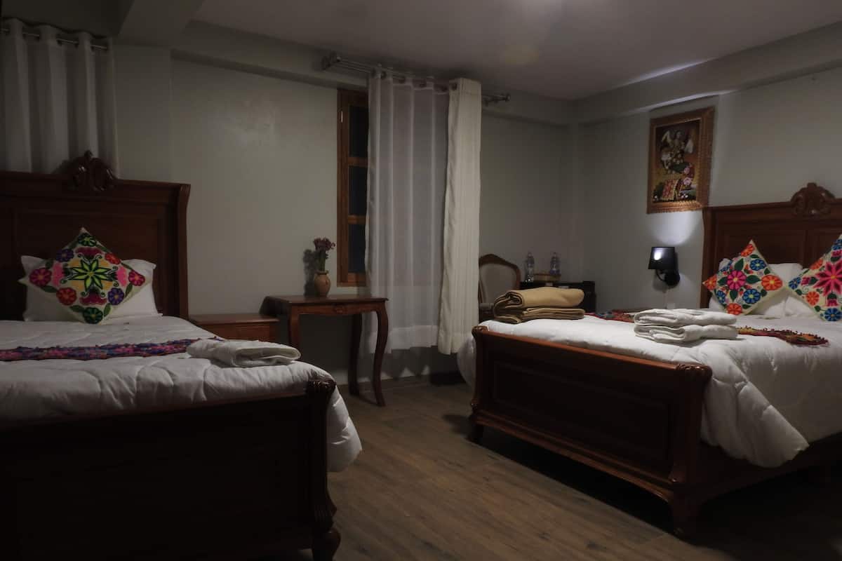 Camera d'albergo con due letti di legno, biancheria bianca, un piccolo tavolo con un vaso e una lampada a parete.
