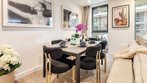 A dining area with a table set for four, surrounded by chairs. There are framed artworks on the walls, a window with a view, and a sofa with a fluffy blanket. - The London Dock HQ - 3 Beds for 6 Guests with AC (London)