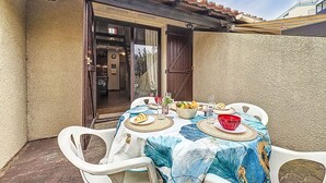 An outdoor patio with a table set for dining, featuring a patterned tablecloth, white plastic chairs, and a variety of food and drink items. - Les Maisons de la Plage by Interhome (Le Grau du Roi)