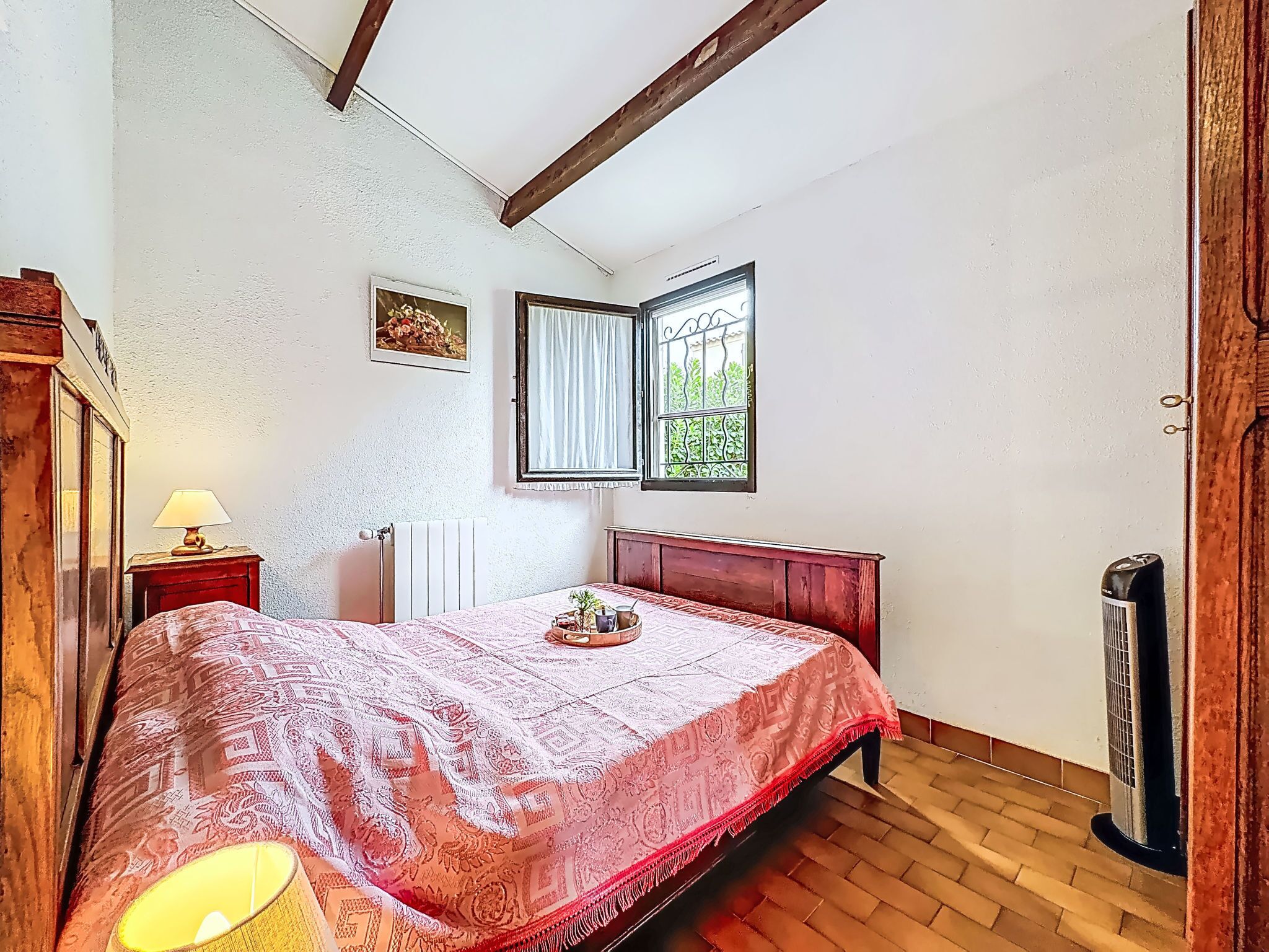 Una camera da letto con un letto, un comodino con una lampada, un termosifone, una finestra con tende e un altro termosifone a pavimento.