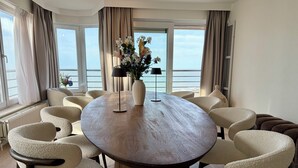 A round table with a vase of flowers, surrounded by chairs, in a room with large windows overlooking the sea.