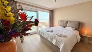 A hotel room with a large bed, a nightstand, a vase of flowers, and a view of the sea through the window.