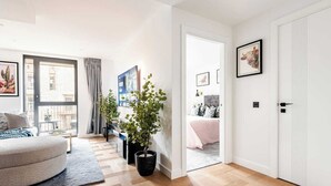 Un appartement moderne comprenant un salon, une chambre et un couloir. On y trouve deux plantes en pot, une grande fenêtre et une porte donnant sur une autre pièce.