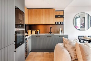 Una cocina moderna con armarios de madera, horno empotrado y pileta. Hay un espejo redondo en la pared y un sofá color beige en la sala.