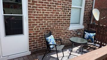 Un patio con muro de ladrillo, puerta corrediza de vidrio, una mesa pequeña con dos sillas y un piso de madera.