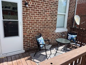 Eine Terrasse mit Backsteinmauer, einer Glastür zum Verschieben, einem kleinen Tisch mit zwei Stühlen und einem Holzboden.