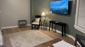 A living room with a television mounted on the wall, a ceiling fan, a chair, a small table with a lamp, and a decorative plant.