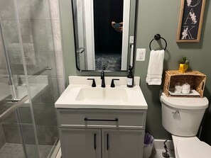 Ein Badezimmer mit einem weißen Waschbecken, einem schwarzen Wasserhahn und einem Spiegel darüber. Eine Toilette mit einem weißen Handtuch an der Wand. Auf dem Toilettentank steht ein kleiner Topfpflanze.
