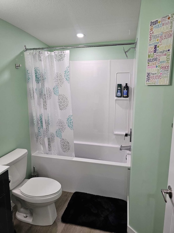 A bathroom with a toilet, bathtub, and shower curtain. There is a small shelf with toiletries. - Brighton B-Suite (Saskatoon)