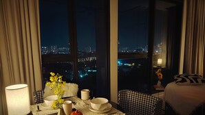 Ein Hotelzimmer mit Esszimmer, gedecktem Tisch für zwei Personen, einer Vase mit gelben Blumen und Blick auf eine nächtliche Stadtlandschaft.