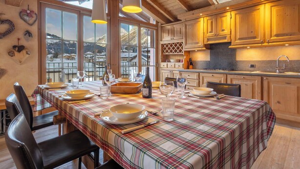 A dining area with a table set for four, wooden cabinets, and a view of a snowy landscape through large windows.