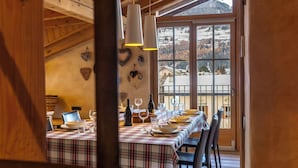 A dining area with a table set for six, wooden beams on the ceiling, and a view of a snowy landscape through large windows.