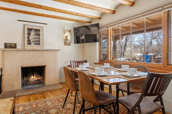 Un comedor con chimenea, vigas de madera, un ventanal con vistas a árboles y un televisor de pantalla plana montado en la pared.