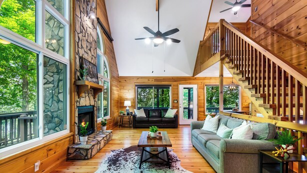 A spacious living room with a stone fireplace, a wooden staircase, and large windows offering a view of the outdoors.