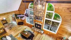 A spacious loft-style room with a stone fireplace, wooden floors, and large windows offering a view of the outdoors.