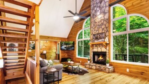 A cozy cabin interior with a stone fireplace, wooden floors, a ceiling fan, and large windows offering a forest view.