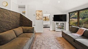 A modern living room with a staircase, a flat-screen TV, and a large window with a view of a forest.