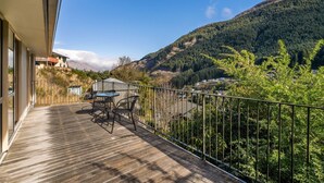 A wooden deck with a table and chairs, overlooking a mountainous landscape with dense forest and a clear sky.