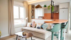 Una sala de estar moderna con un sofá, una mesa de centro y sillas. Hay una zona de cocina con un refrigerador y un lavamanos.