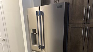 Un refrigerador de acero inoxidable con dispensador de agua y un cajón congelador, situado en una cocina con armarios de madera oscura y suelo de baldosas de color claro.