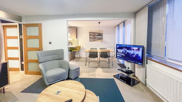 A modern living room with a grey armchair, a wooden coffee table, a TV, and a dining area with a table and chairs.