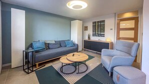A modern living room with a grey sofa, a blue armchair, a small wooden coffee table, and a TV stand. - Entire Birmingham Home | Sleeps 4, Parking & Garden (Lozells)