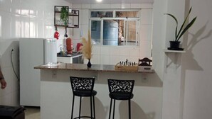 A compact kitchen with white tiles, a small island with two black bar stools, a refrigerator, and a potted plant. - Mumu's Corner (Ouro Preto)