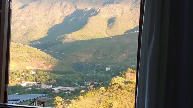 A room with a large arched window offering a view of a valley with mountains and a small settlement. - Mumu's Corner (Ouro Preto)