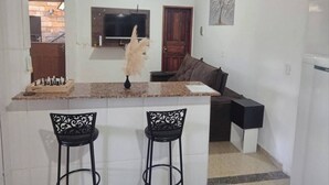 A compact kitchen with a central island, two bar stools, a television above the island, and a door leading to a brick area. - Mumu's Corner (Ouro Preto)