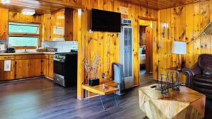A cabin interior with wooden walls, a kitchen area, a living room with a sofa, and a TV.