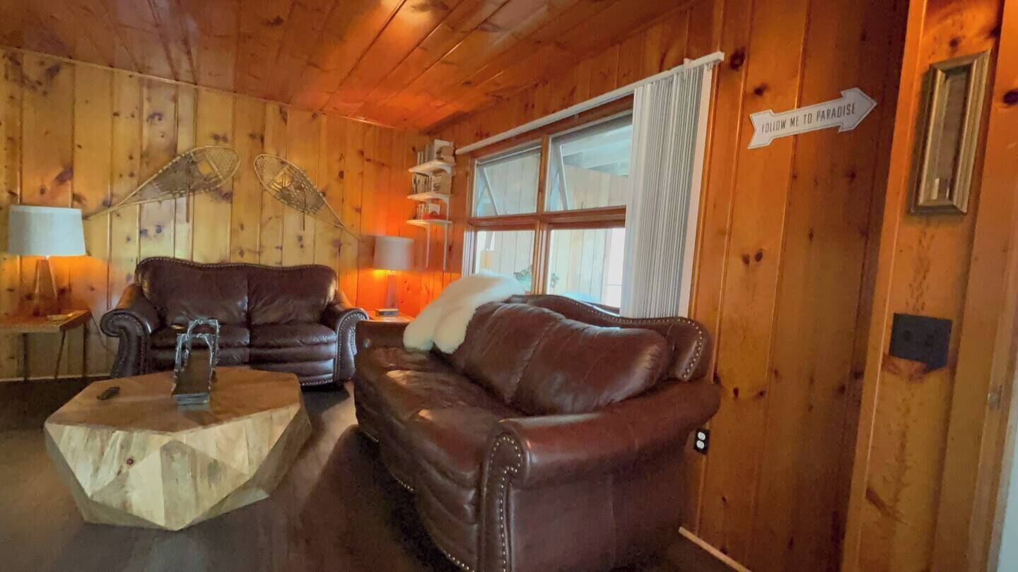 Interior de una cabaña de madera con un sofá de cuero marrón, una mesa de centro de madera y una ventana con cortinas.