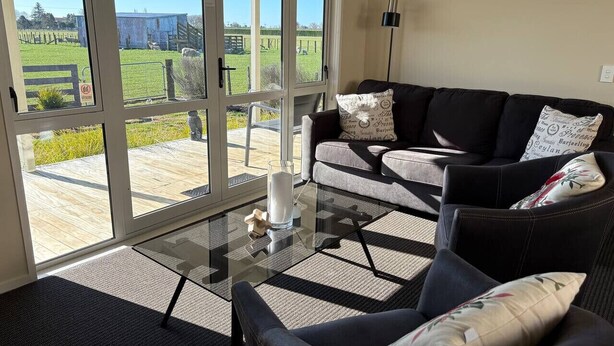 A modern living room with a glass coffee table, two sofas, and a view of a green landscape through large sliding glass doors.
