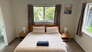Ein ordentlich bezogenes Bett mit einem gefalteten Handtuch, zwei Nachttische mit Lampen, ein Fenster mit Vorhängen und ein Bild an der Wand.