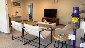 Ein Wohnzimmer mit einer Couch, zwei Sesseln, einem Couchtisch, einem Fernseher und einem Staubsauger.
