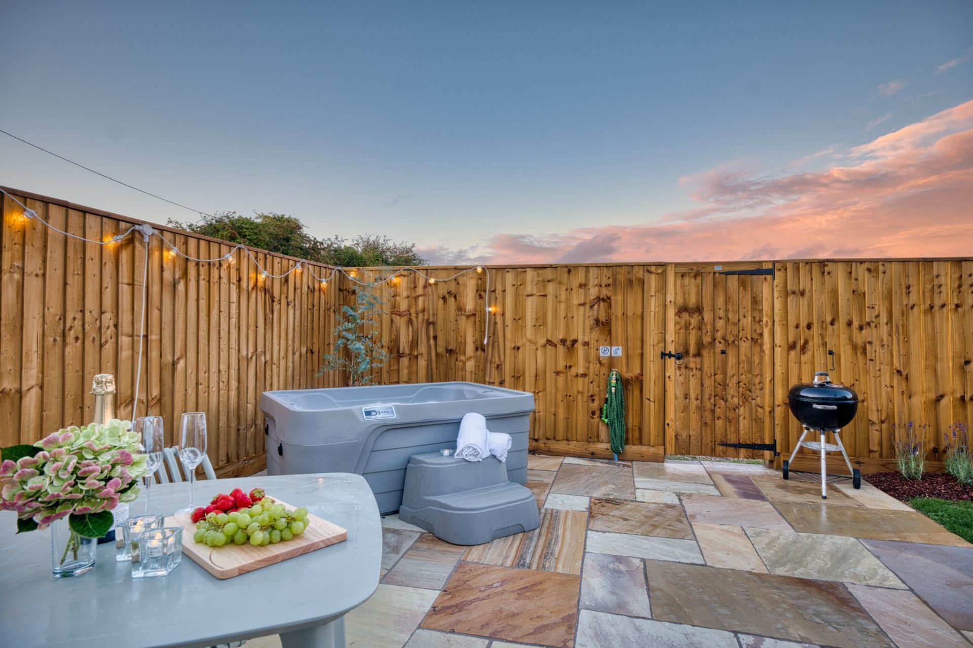 Patio trasero con jacuzzi, una parrilla de barbacoa y una mesa con una bandeja de fruta y vasos.