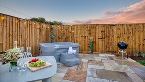 Patio trasero con jacuzzi, una parrilla de barbacoa y una mesa con una bandeja de fruta y vasos.