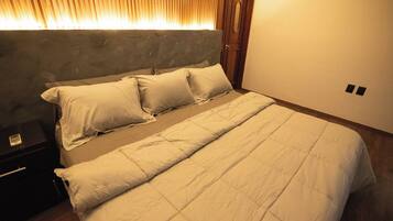 A neatly made bed with a grey headboard and white bedding in a room with wooden flooring and a wooden door.