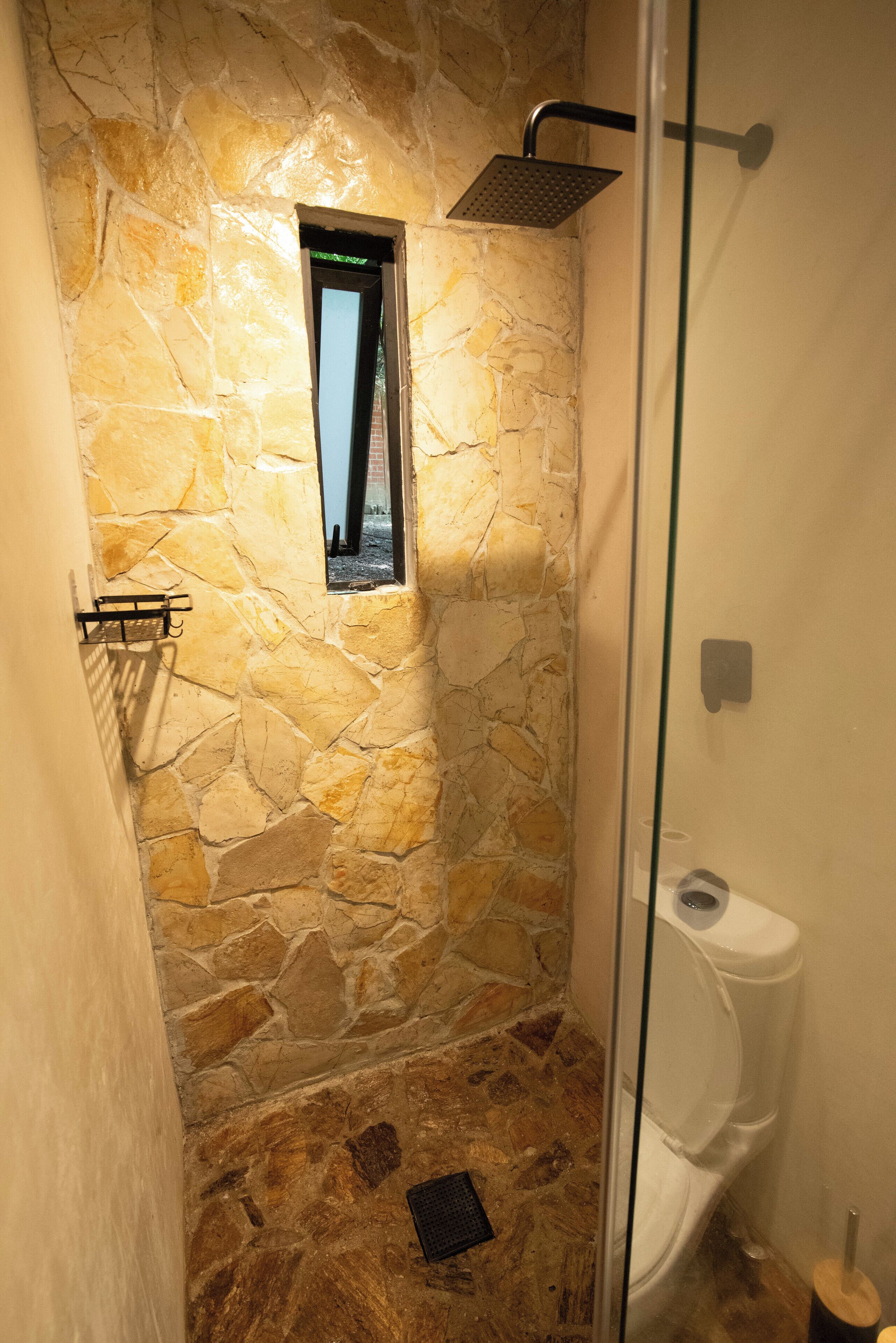 Dutxa amb parets de pedra, una porta de vidre, una finestra petita i un vàter blanc.