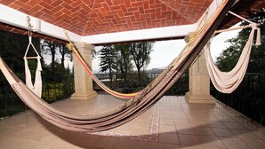 Un hamac est suspendu entre des colonnes dans un espace extérieur abrité, doté d’un sol carrelé et offrant une vue sur les arbres et un paysage lointain.