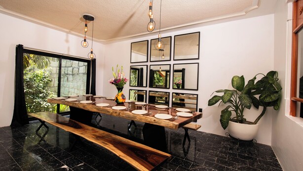 Un espace repas avec une longue table en bois, dressée pour accueillir plusieurs personnes, entouré d’un sol en marbre noir et agrémenté d’une grande fenêtre donnant sur un paysage verdoyant.