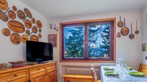 Ein Essbereich mit einem Tisch für vier Personen, einer Holzkommode mit Fernseher und einem Fenster mit Blick auf Bäume.