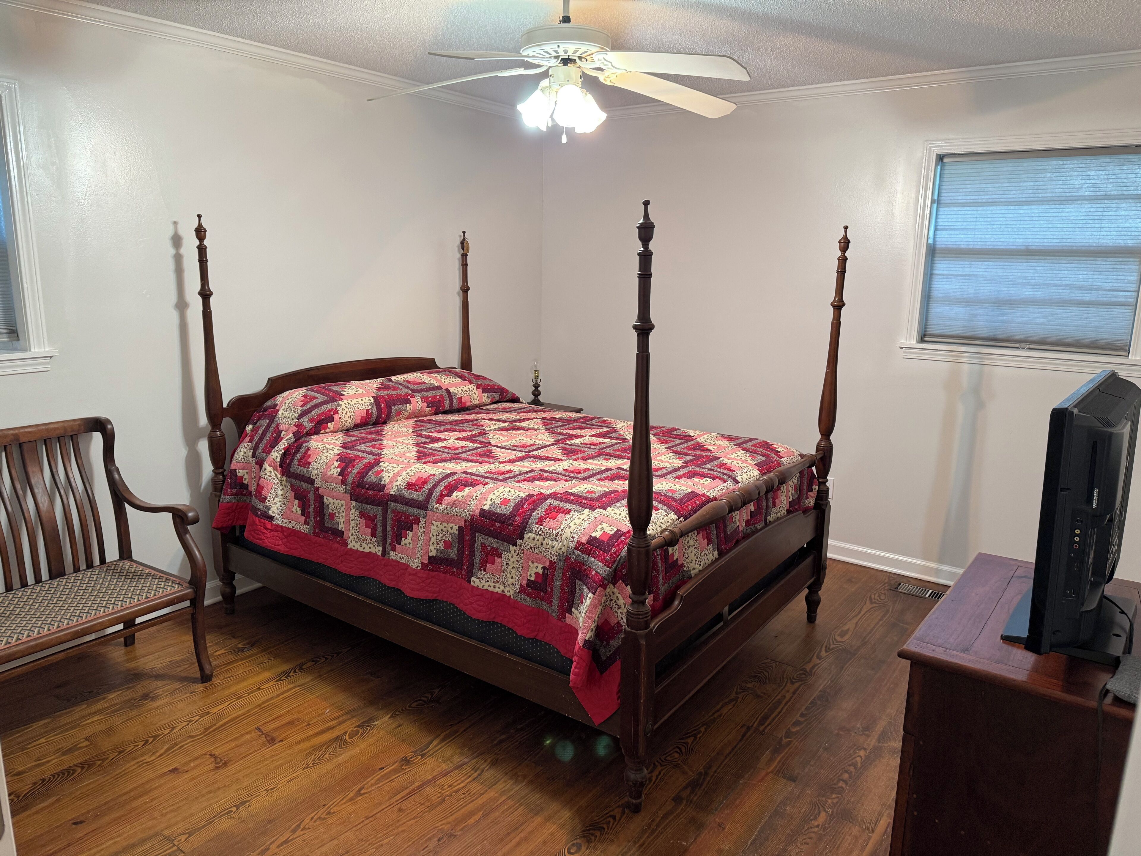 A four-poster bed with a patterned quilt, a wooden chair, a television on a wooden stand, and a ceiling fan.