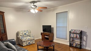 A living room with a leather sofa, a patterned armchair, a wooden coffee table, a television on a wooden cabinet, and a bookshelf filled with DVDs. - Charming, newly renovated 3-bedroom house just minutes from downtown Oxford. (Oxford)