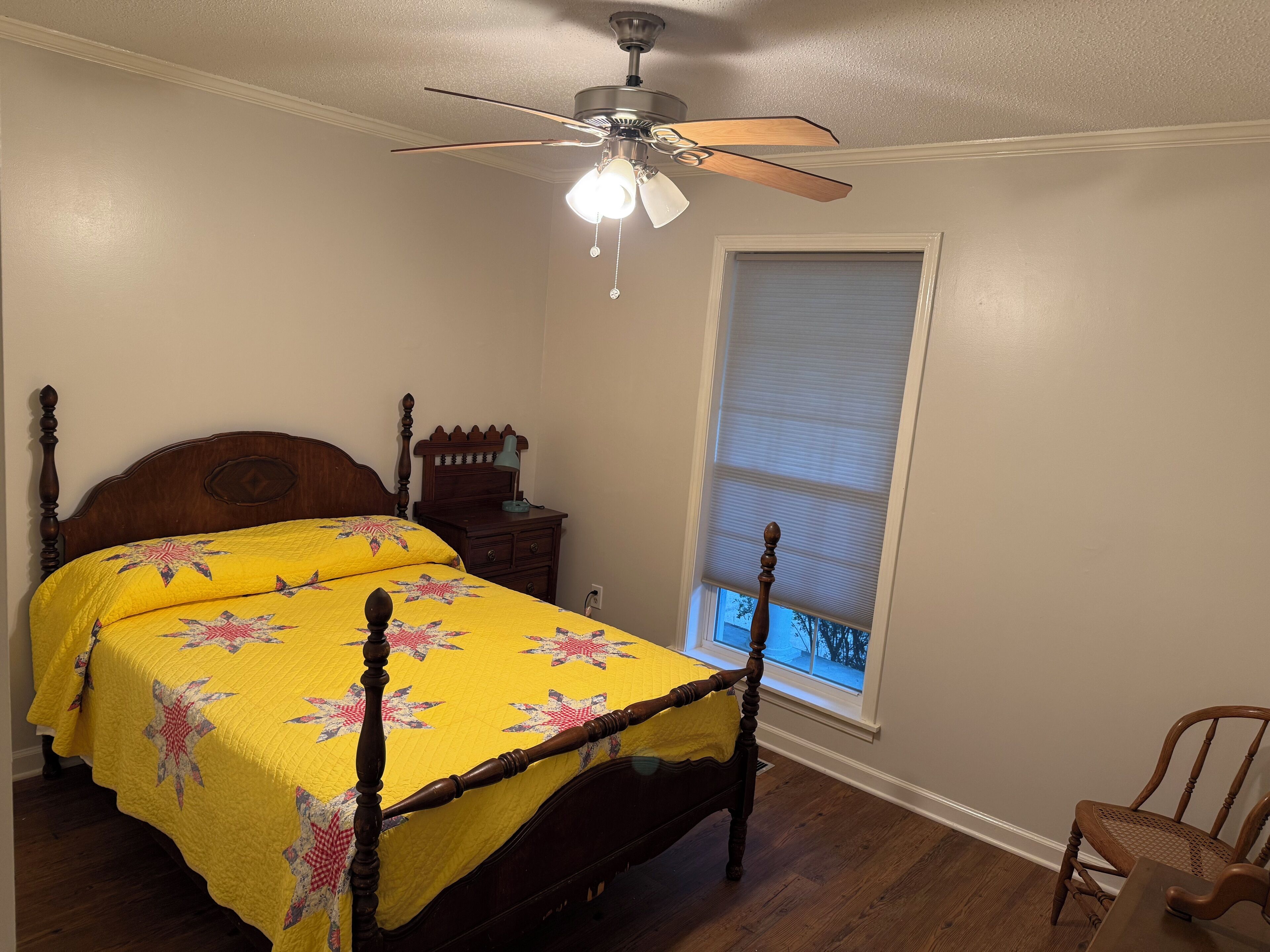 A bedroom with a wooden bed, a nightstand, a desk, and a window with blinds.
