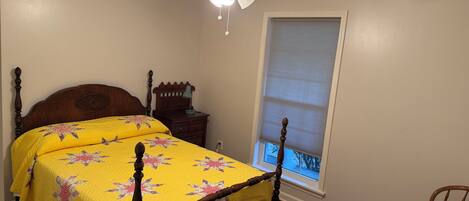 A bedroom with a wooden bed, a nightstand, a desk, and a window with blinds.